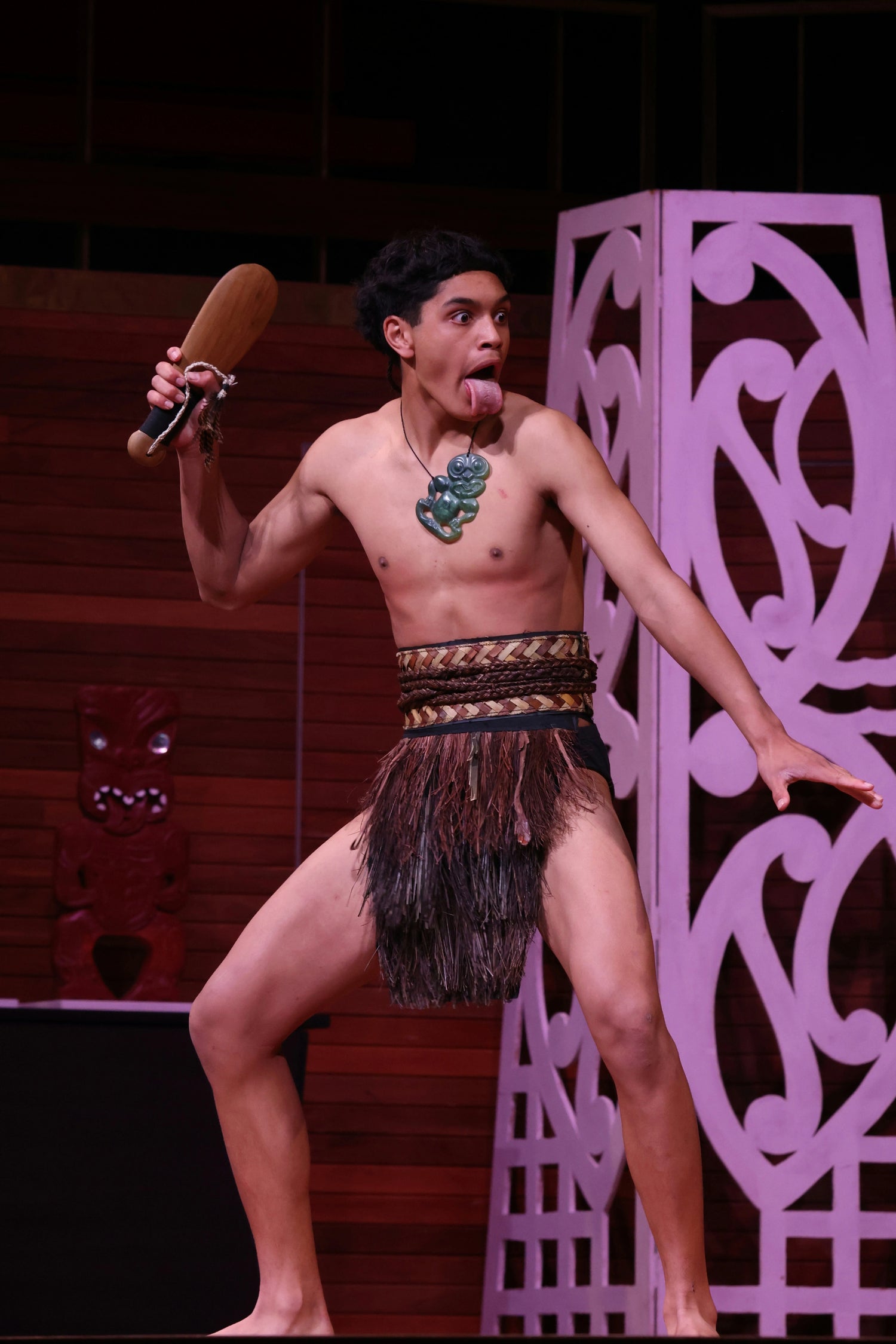 Person in traditional attire holding a wooden object on a stage with decorative elements.